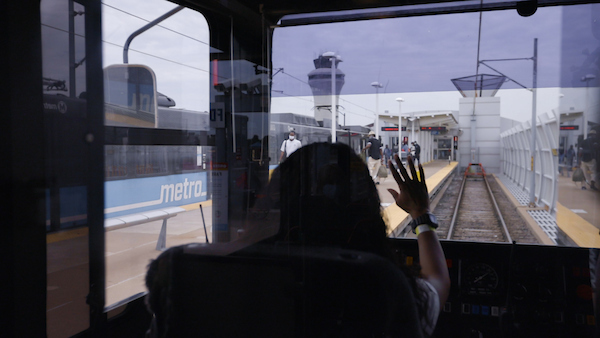 Interior shot of a MetroLink operator waving to riders on the platform of the Lambert Airport Terminal #1 MetroLink station. The platform and airport control tower can be seen through the windshield.